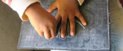 A kindergarten child traces her fingers with chalk on a blackboard to develop motor skills during a creative lesson taught by Projects Abroad Childcare volunteers in Madagascar. A child outlines her hand with chalk during a skills development lesson with a volunteer working with children in Madagascar.